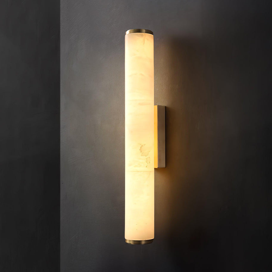 Wall lamp with a long pale veined stone-look tube and a gold-toned bracket, lit, on a dark grey wall.