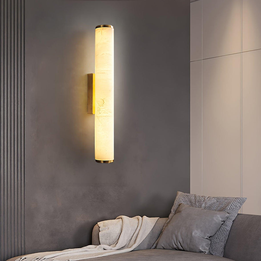 Wall lamp with a long pale veined alabaster tube and a brass-coloured bracket lit above a bed.