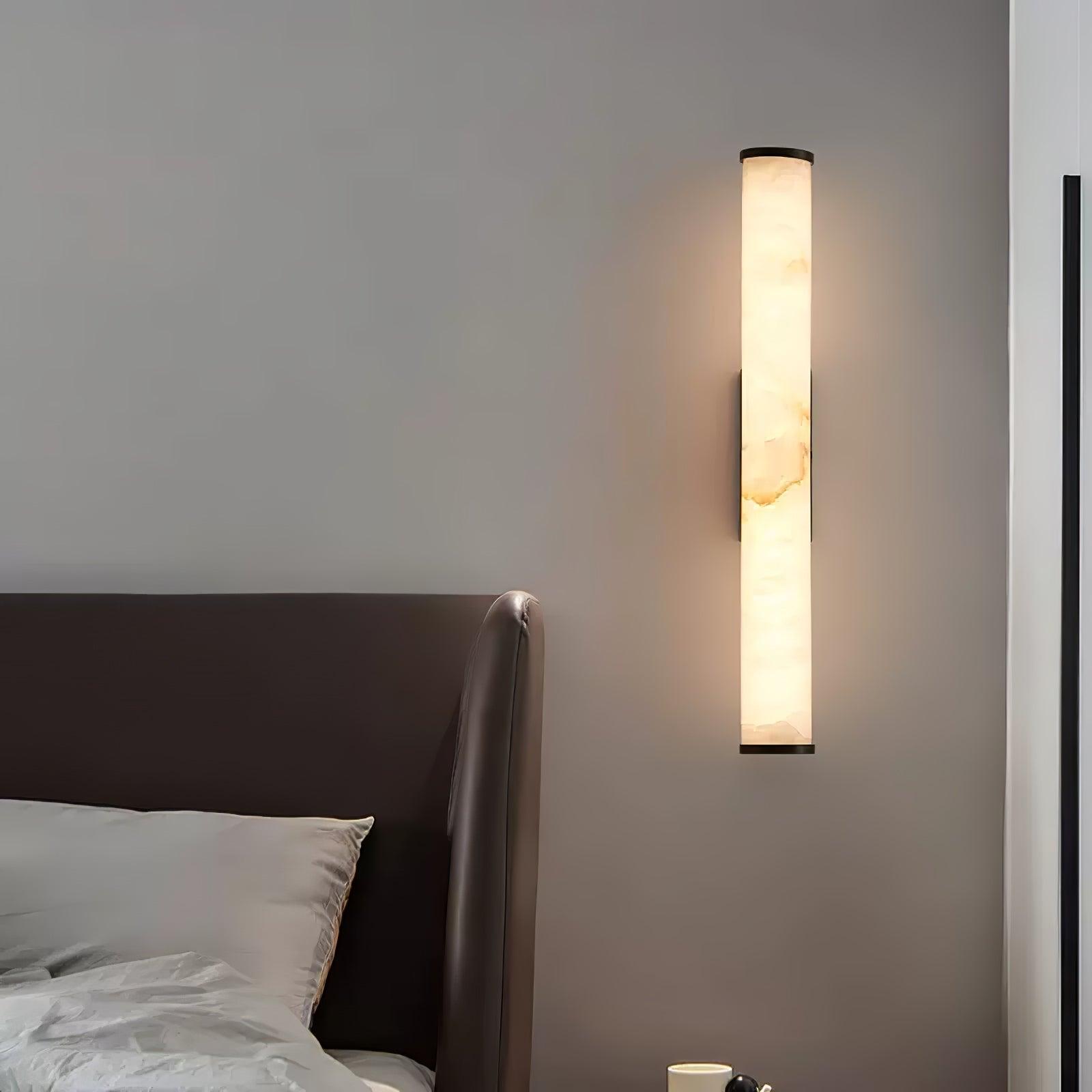 Wall lamp with a long pale veined stone-look tube and a dark bracket, lit, on a wall beside a leather headboard.