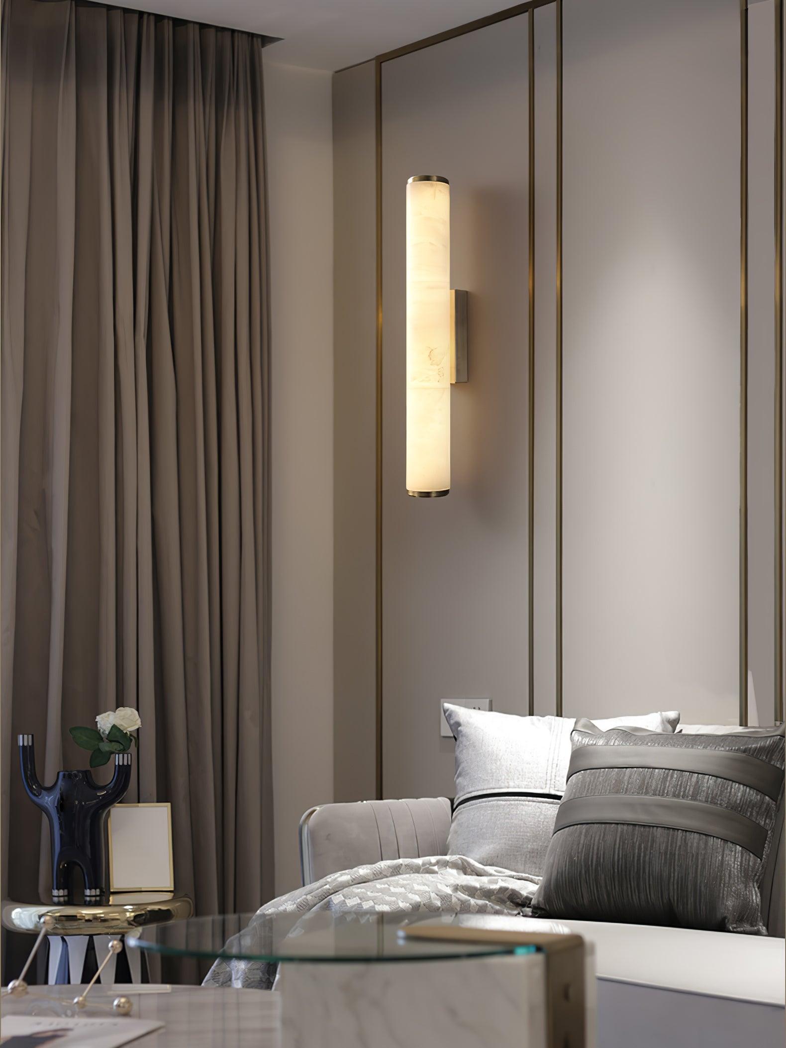Wall lamp with a long pale veined alabaster tube and a black bracket lit beside a mirrored panel and curtain.
