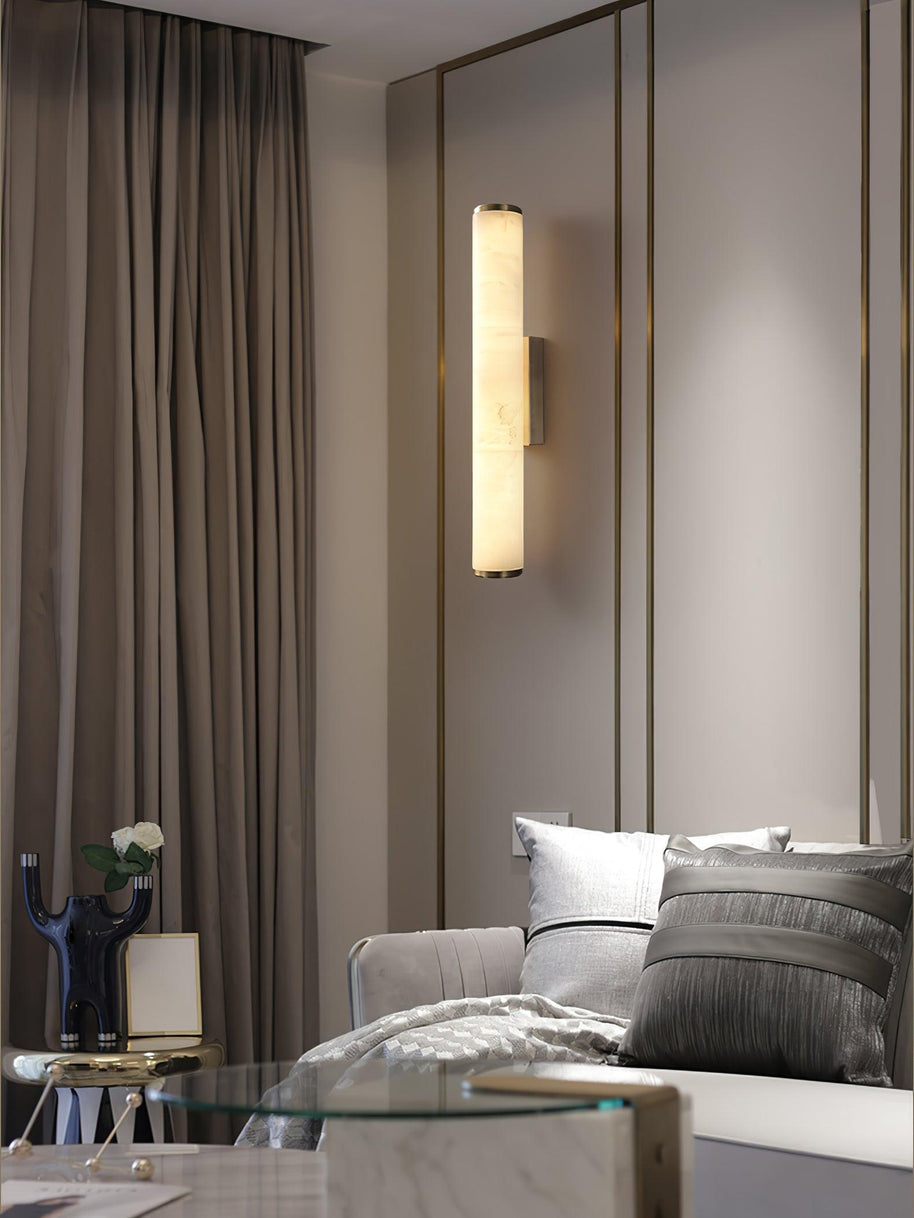 Wall lamp with a long pale veined alabaster tube and a black bracket lit beside a mirrored panel and curtain.