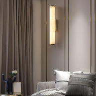 Wall lamp with a long pale veined alabaster tube and a black bracket lit beside a mirrored panel and curtain.