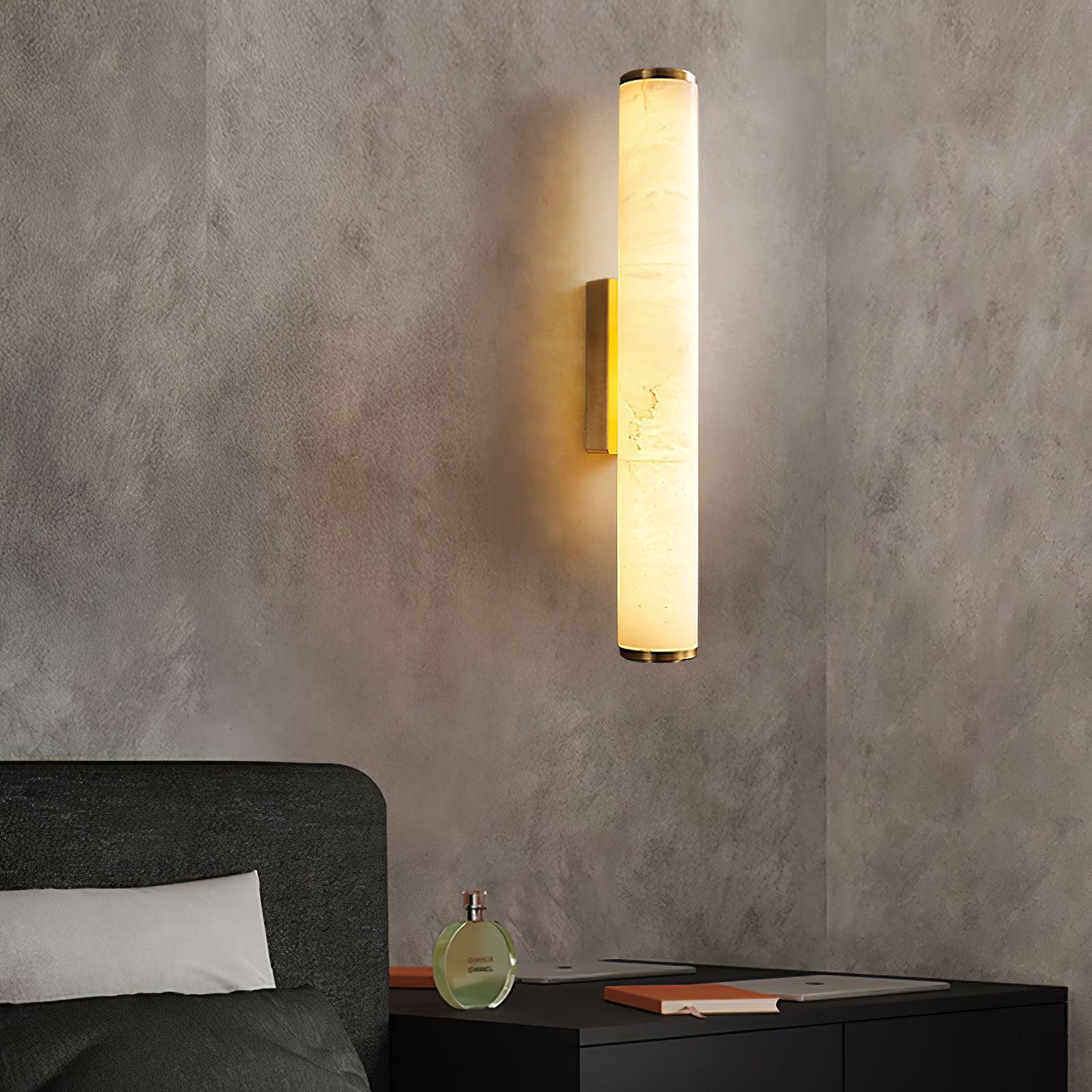 Wall lamp with a long pale veined alabaster tube and a brass-coloured bracket lit above a bedside table.