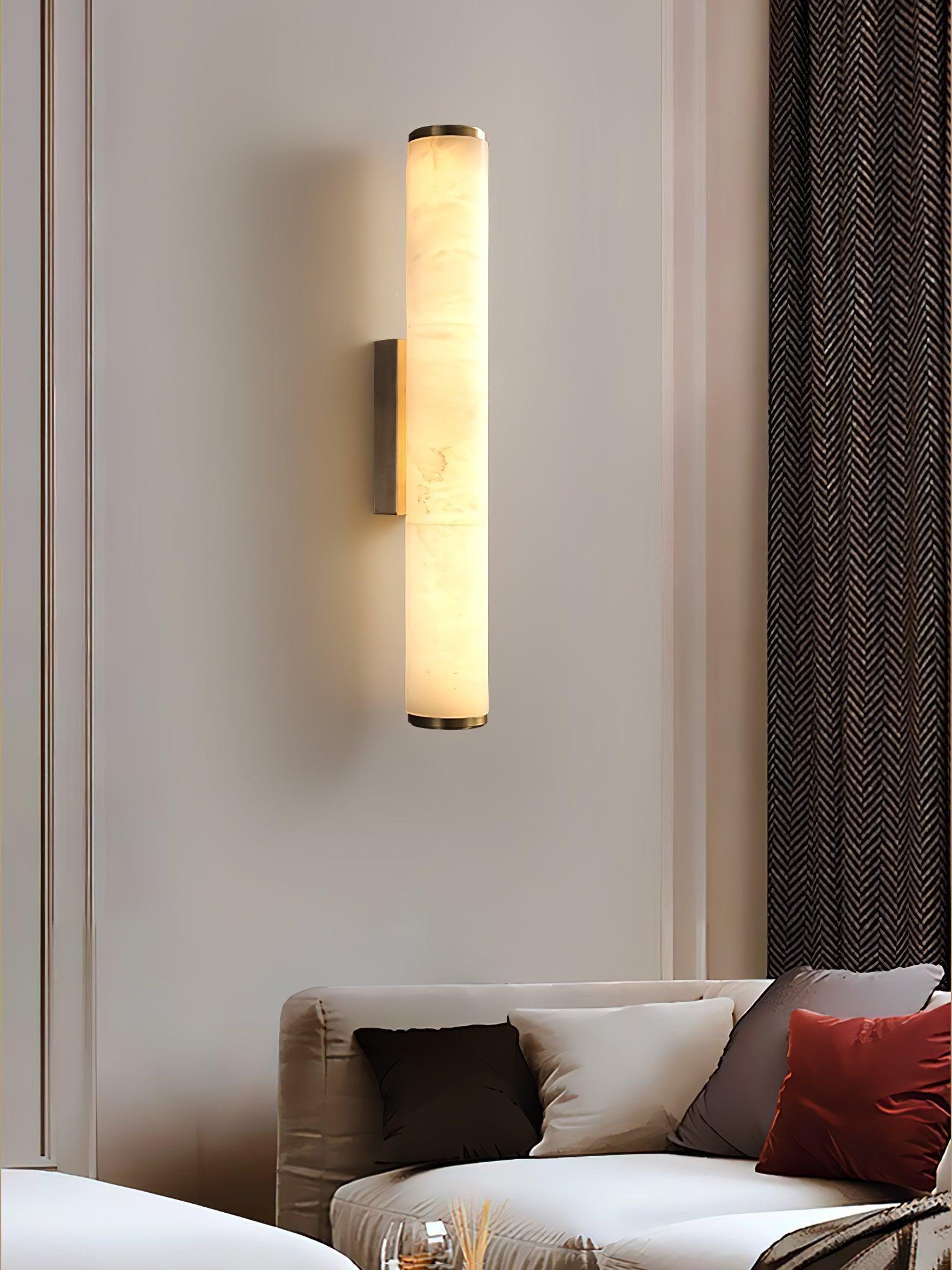 Wall lamp with a long pale veined alabaster tube and a black bracket lit beside a slatted wall panel.