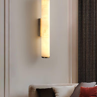 Wall lamp with a long pale veined alabaster tube and a black bracket lit beside a slatted wall panel.