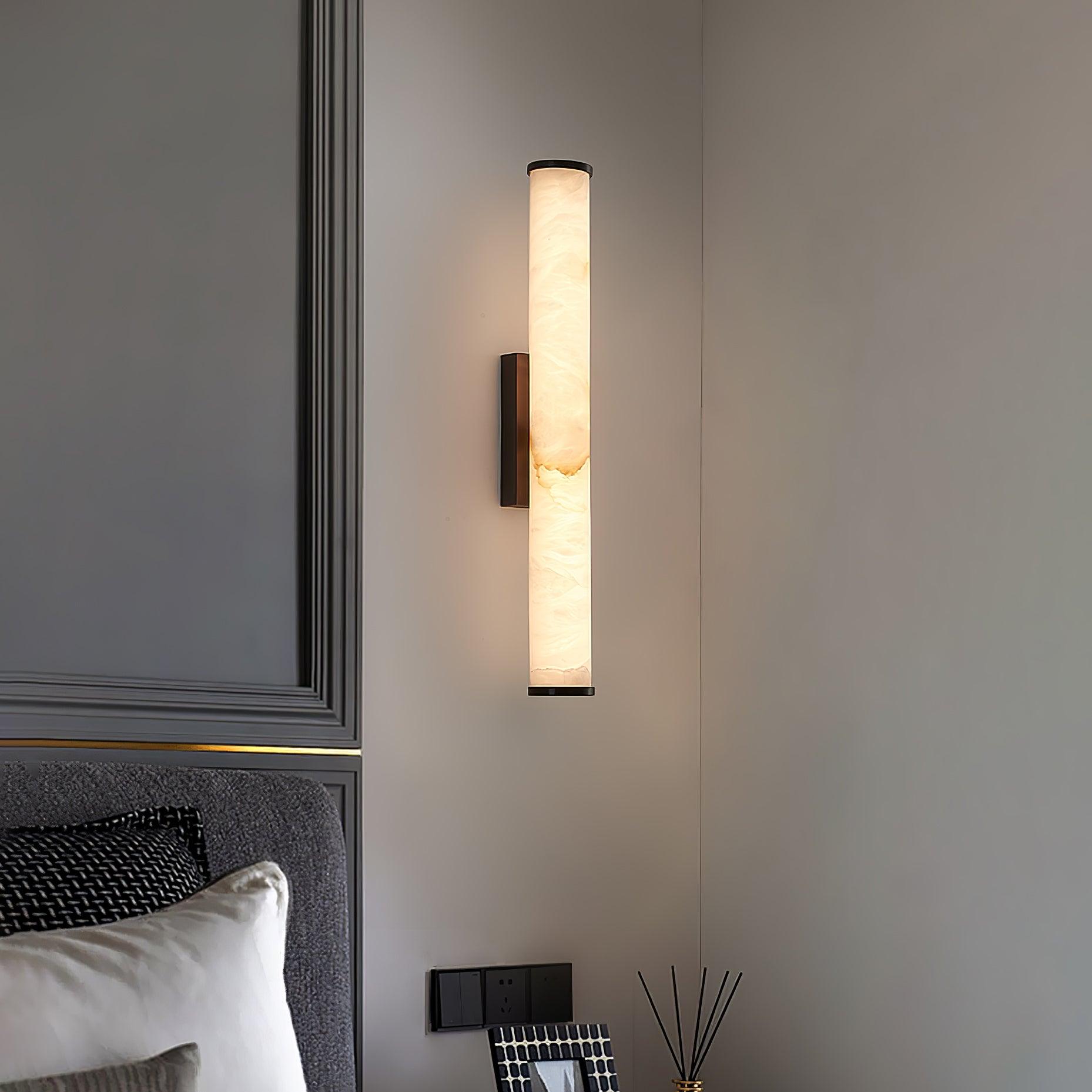 Wall lamp with a long pale veined alabaster tube and a black bracket lit on a panelled wall above a bed.