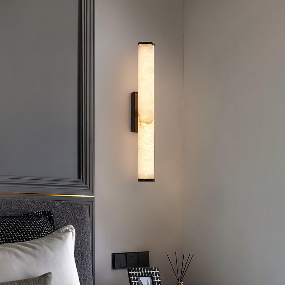 Wall lamp with a long pale veined alabaster tube and a black bracket lit on a panelled wall above a bed.