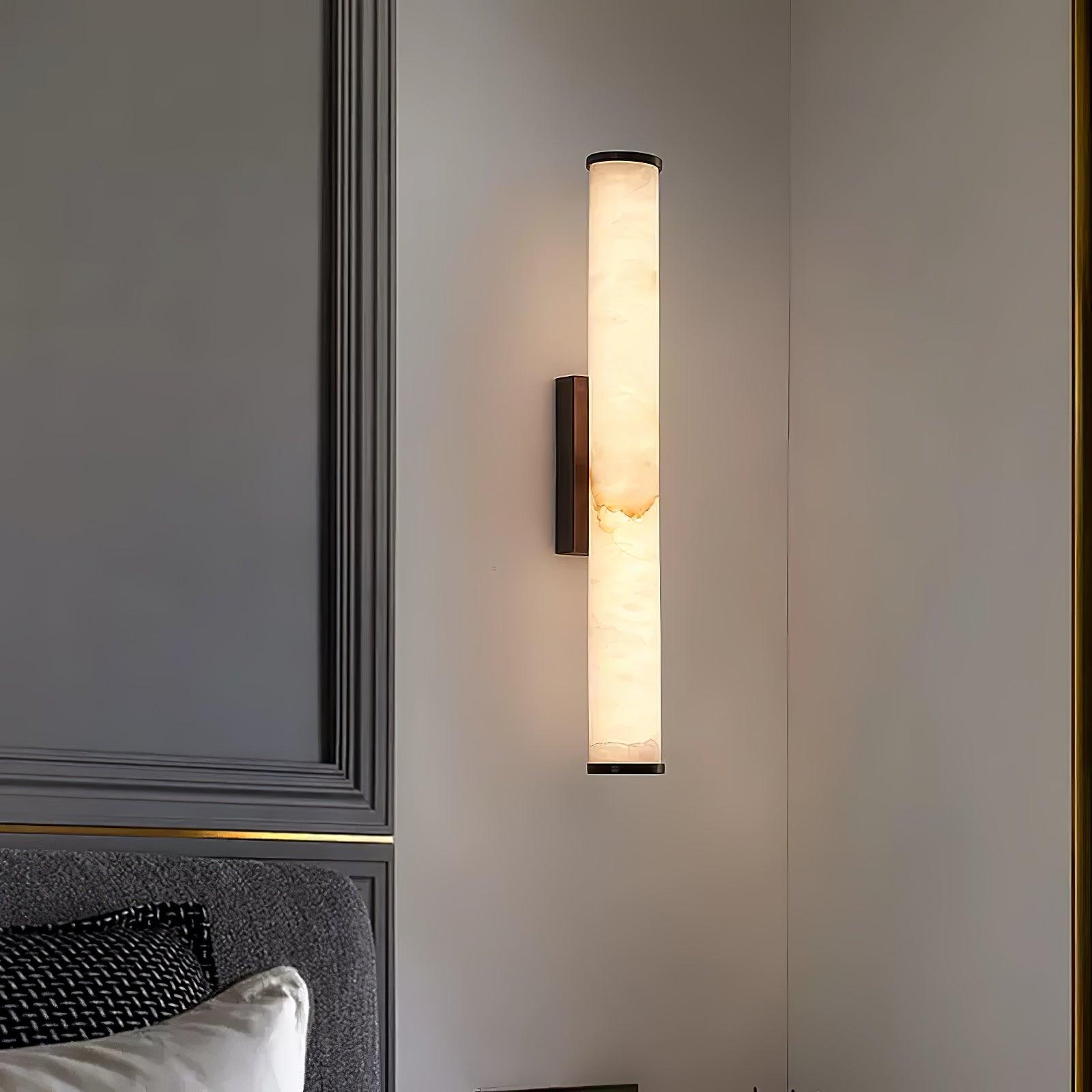 Wall lamp with a long pale veined stone-look tube and a dark bracket, lit, on a panelled wall.