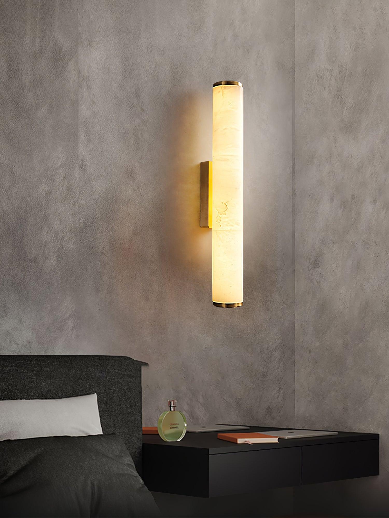 Wall lamp with a long pale veined alabaster tube and a brass-coloured bracket lit on a textured plaster wall above a bed.