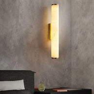 Wall lamp with a long pale veined alabaster tube and a brass-coloured bracket lit on a textured plaster wall above a bed.