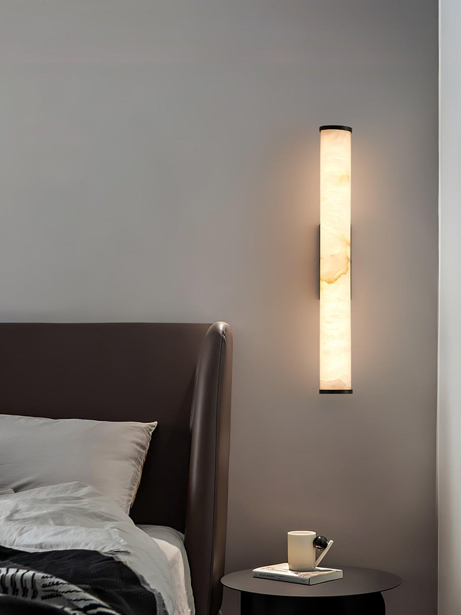 Wall lamp with a long pale veined alabaster tube and a black bracket lit beside a bed.