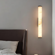 Wall lamp with a long pale veined alabaster tube and a black bracket lit beside a bed.