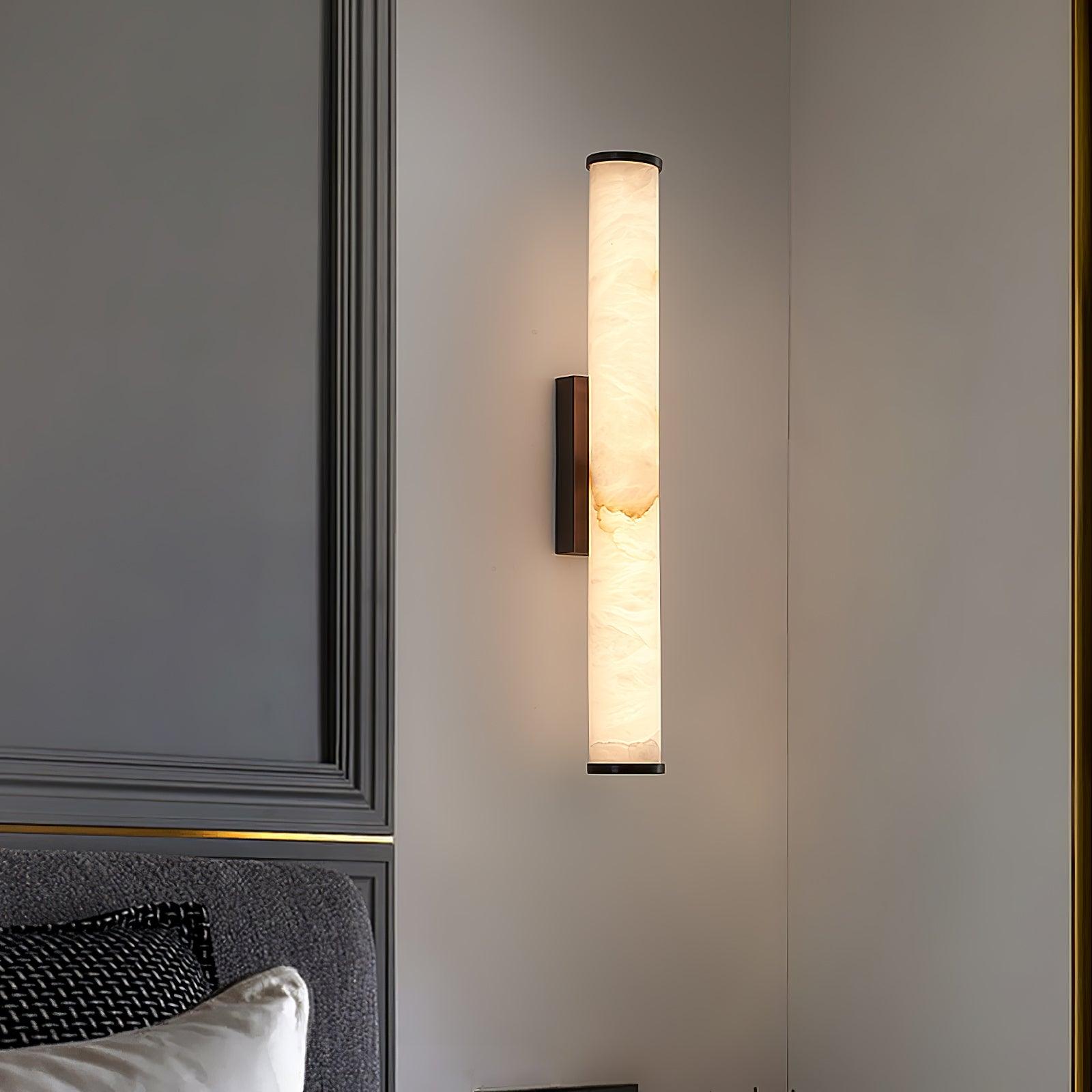 Wall lamp with a long pale veined stone-look tube and a dark bracket, lit, on a panelled wall beside a headboard.