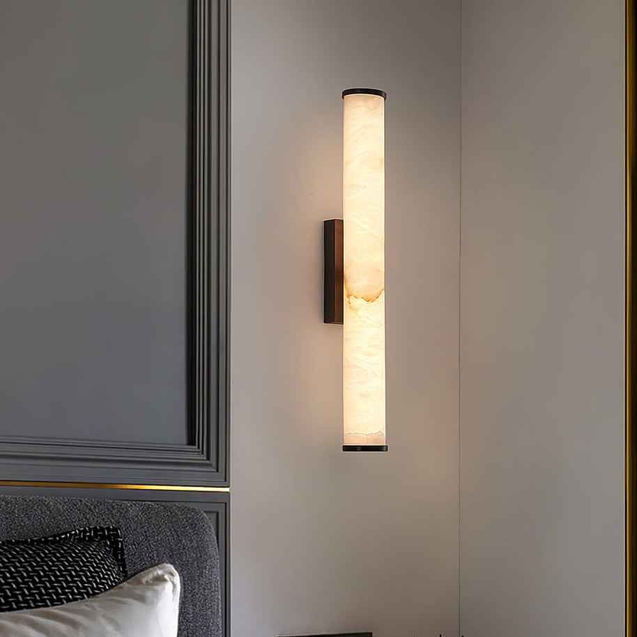 Wall lamp with a long pale veined stone-look tube and a dark bracket, lit, on a panelled wall beside a headboard.