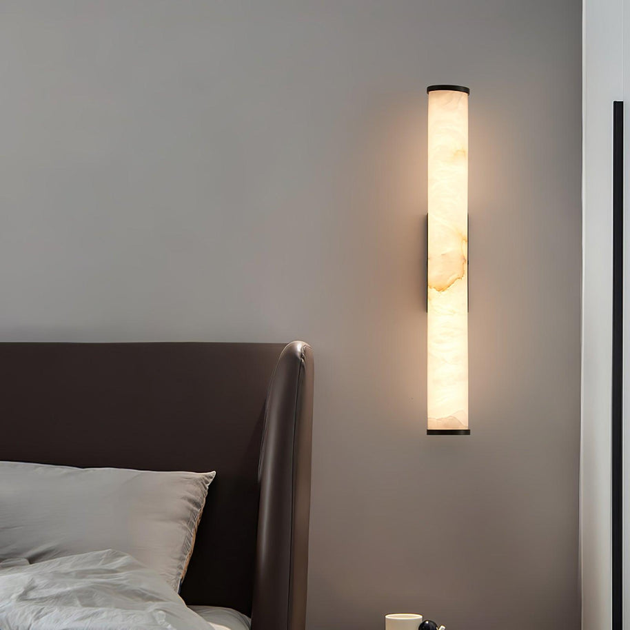 Wall lamp with a long pale veined stone-look tube and a dark bracket, lit, above a bed with a cup on the side table.