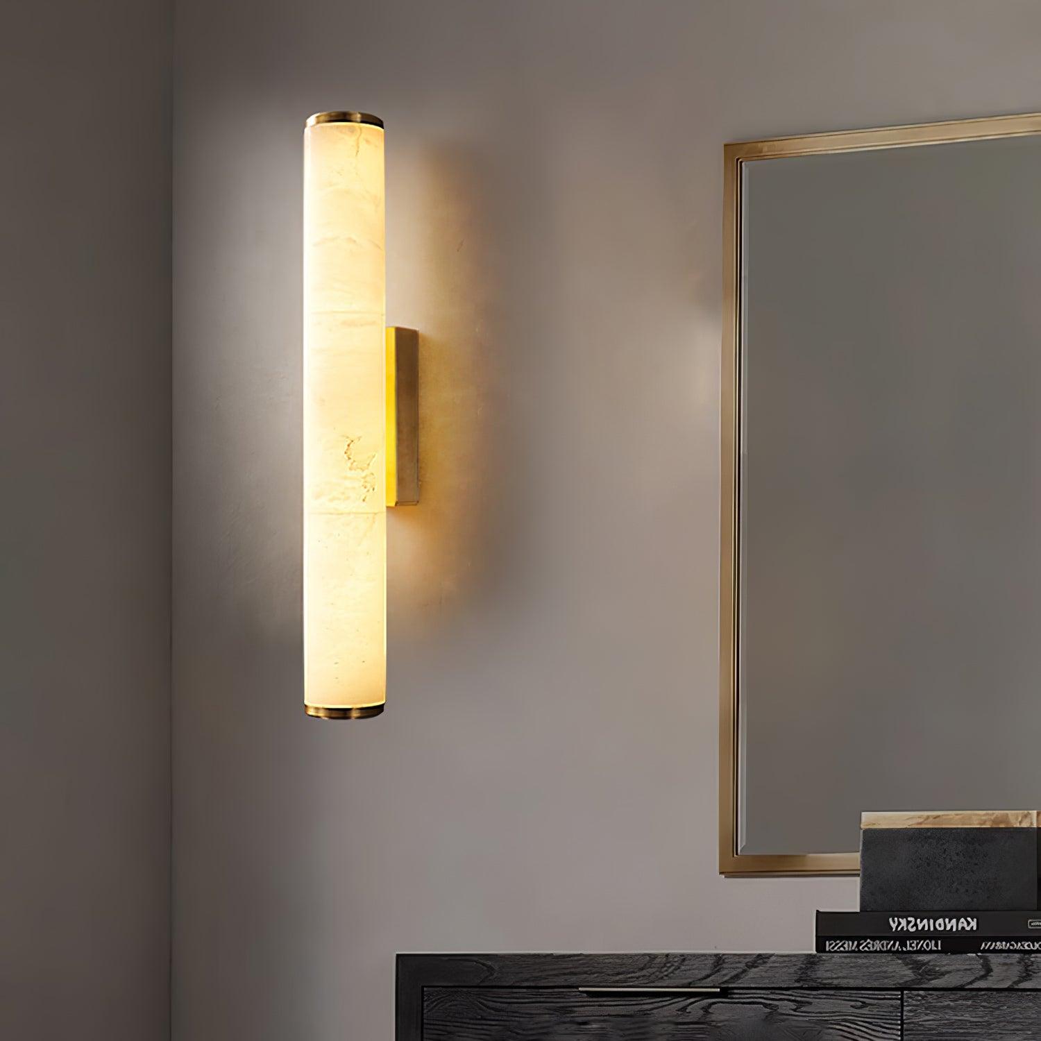 Wall lamp with a long pale veined alabaster tube and a brass-coloured bracket lit on a plaster wall beside a mirror.