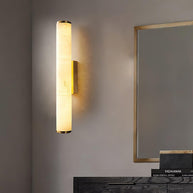Wall lamp with a long pale veined alabaster tube and a brass-coloured bracket lit on a plaster wall beside a mirror.