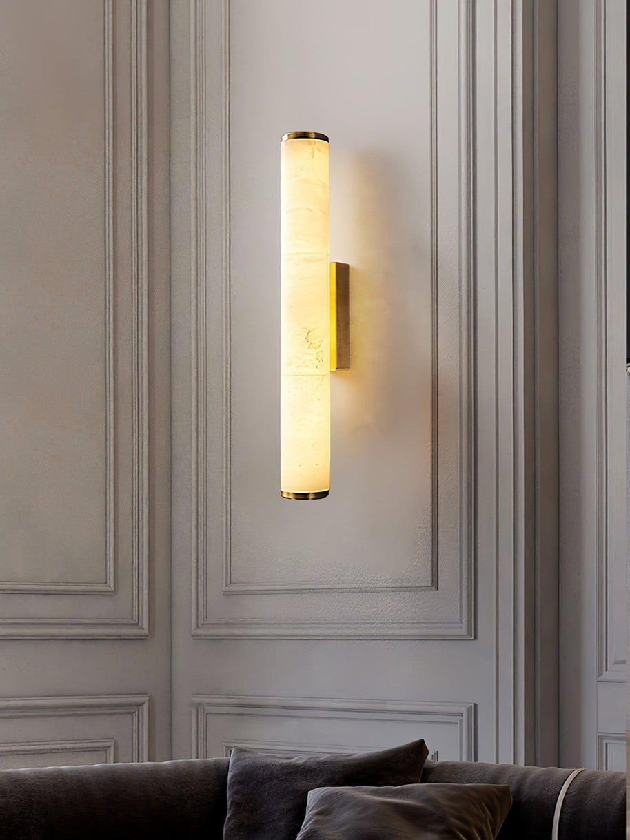 Wall lamp with a long pale veined alabaster tube and a brass-coloured bracket lit on a panelled wall above a sofa.