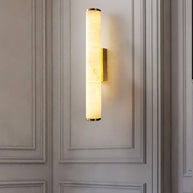 Wall lamp with a long pale veined alabaster tube and a brass-coloured bracket lit on a panelled wall above a sofa.