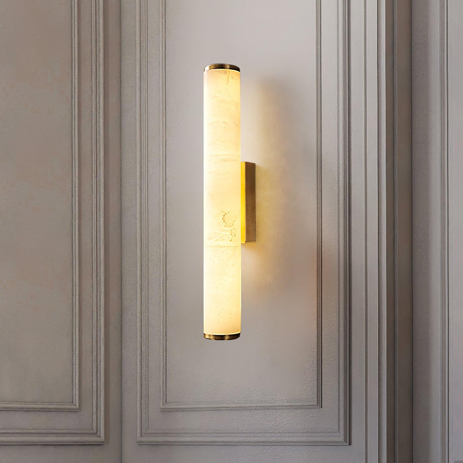Wall lamp with a long pale veined alabaster tube and a brass-coloured bracket lit on a panelled wall.