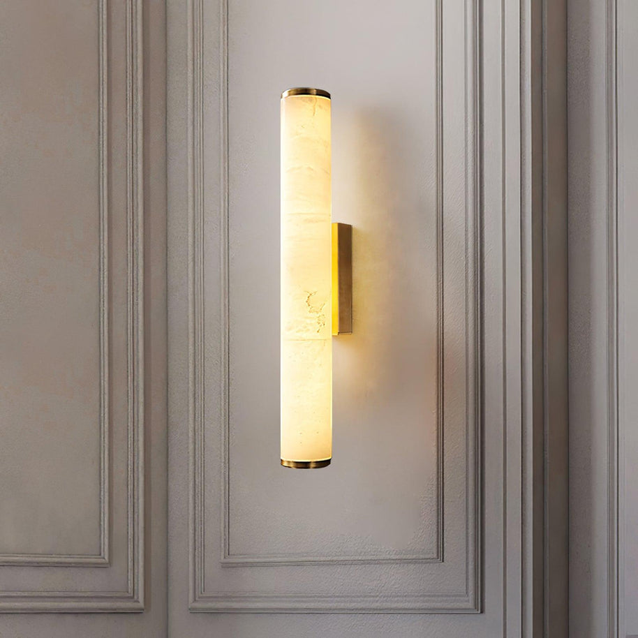 Wall lamp with a long pale veined alabaster tube and a brass-coloured bracket lit on a panelled wall.