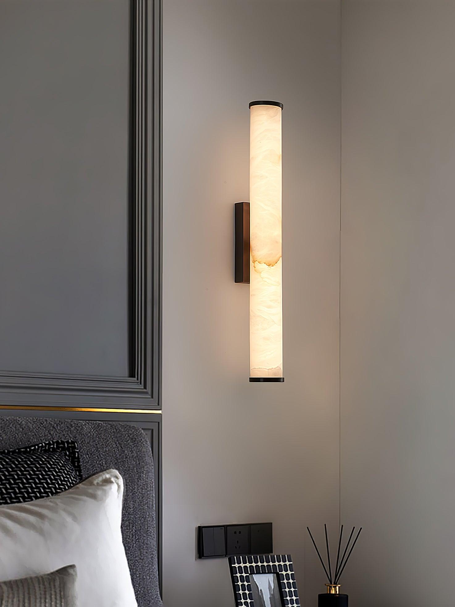 Wall lamp with a long pale veined alabaster tube and a black bracket lit on a panelled wall above a bed.