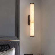 Wall lamp with a long pale veined alabaster tube and a black bracket lit on a panelled wall above a bed.