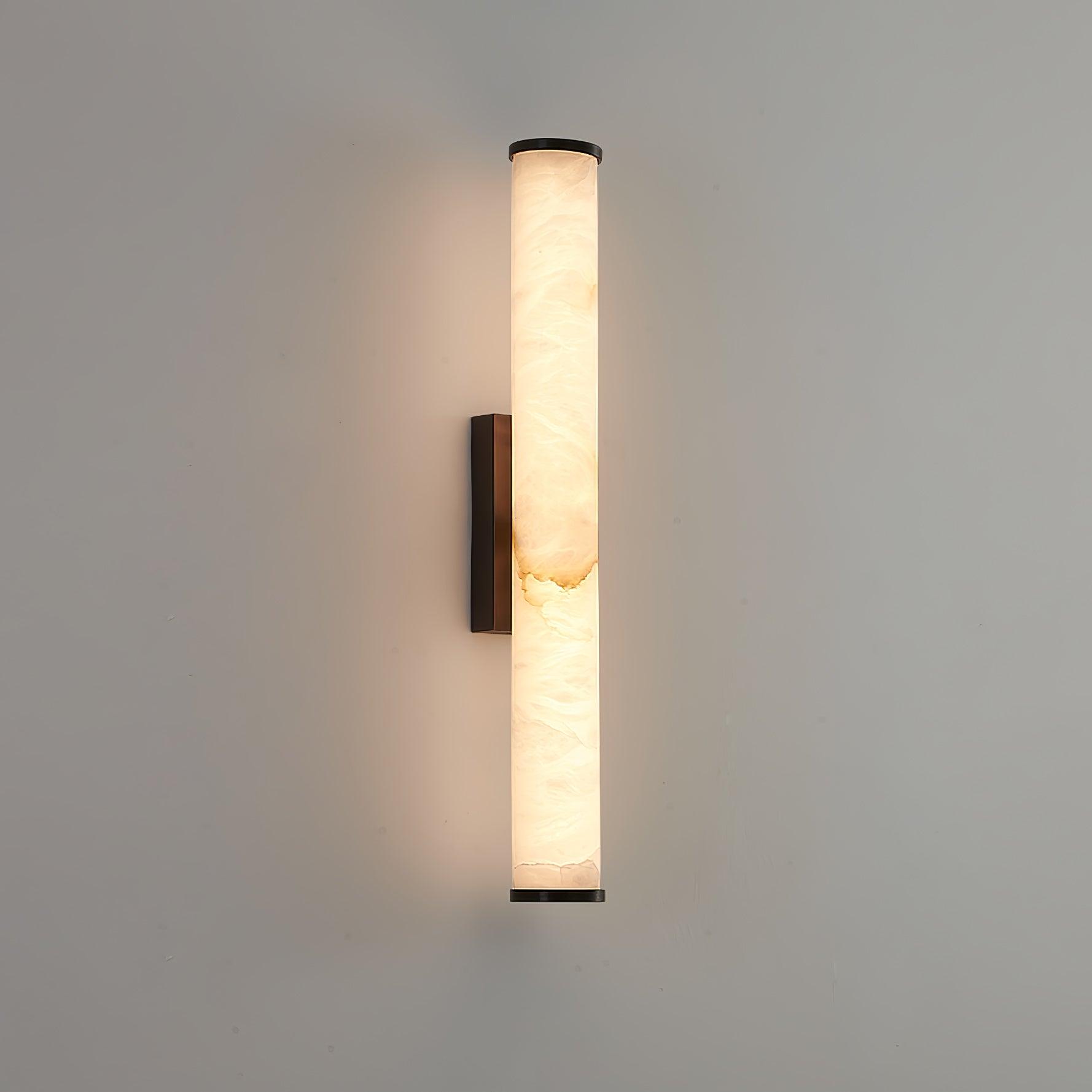 Wall lamp with a long pale veined stone-look tube and a dark bracket, lit, on a grey wall.