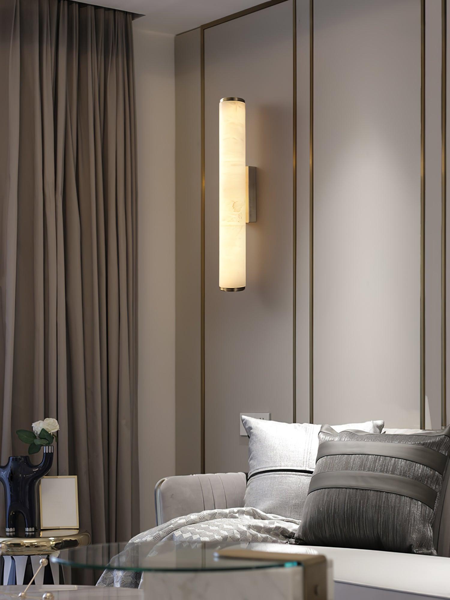 Wall lamp with a long pale veined alabaster tube and a black bracket lit beside a mirrored wall and curtain.