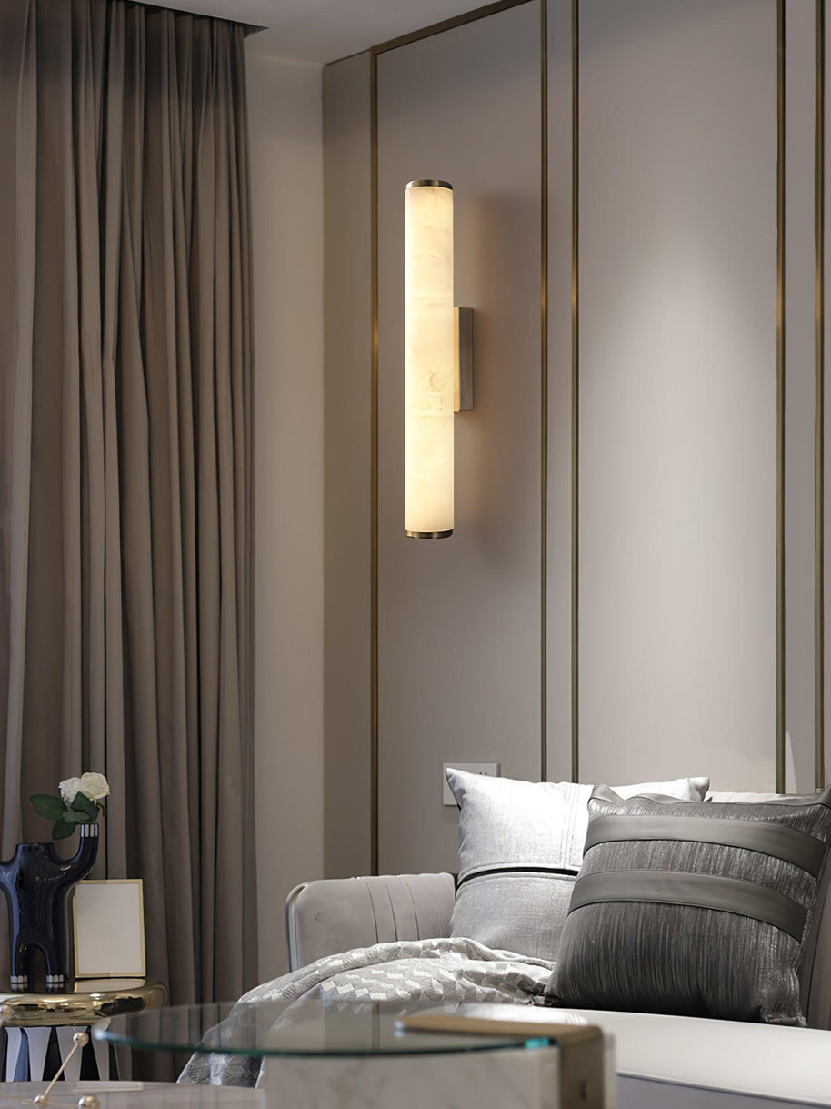Wall lamp with a long pale veined alabaster tube and a black bracket lit beside a mirrored wall and curtain.