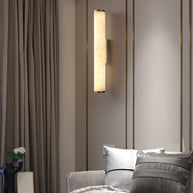 Wall lamp with a long pale veined alabaster tube and a black bracket lit beside a mirrored wall and curtain.