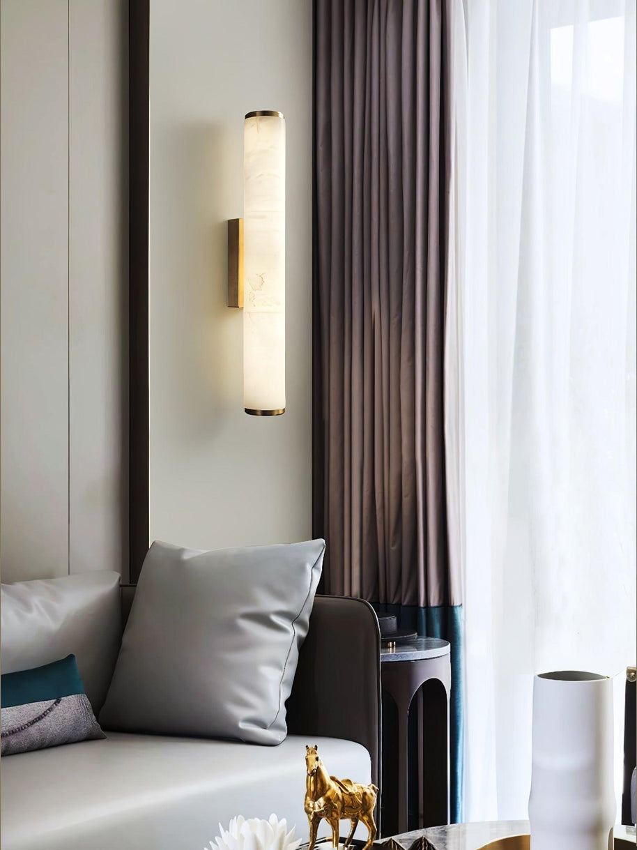 Wall lamp with a long pale veined alabaster tube and a black bracket lit beside a curtain.