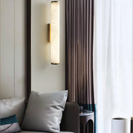 Wall lamp with a long pale veined alabaster tube and a black bracket lit beside a curtain.