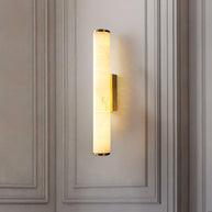 Wall lamp with a long pale veined alabaster tube and a brass-coloured bracket lit on a panelled wall.