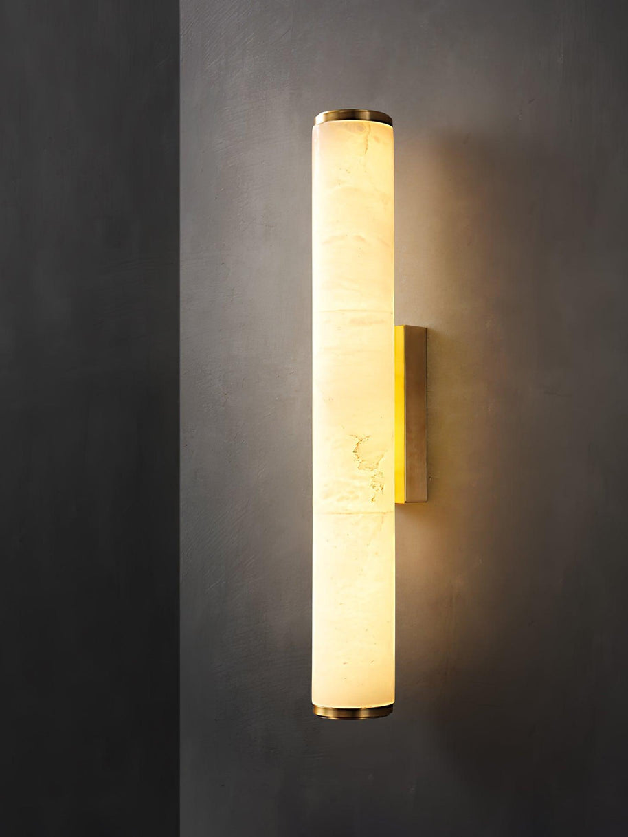 Wall lamp with a long pale veined alabaster tube and a brass-coloured bracket lit on a dark grey wall.