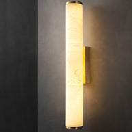 Wall lamp with a long pale veined alabaster tube and a brass-coloured bracket lit on a dark grey wall.