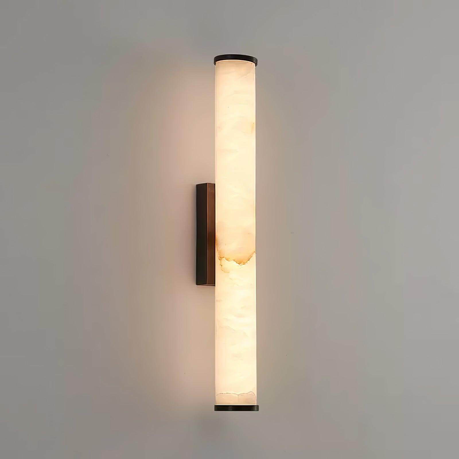 Wall lamp with a long pale veined alabaster tube and a black bracket lit on a plain pale wall.