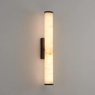Wall lamp with a long pale veined alabaster tube and a black bracket lit on a plain pale wall.