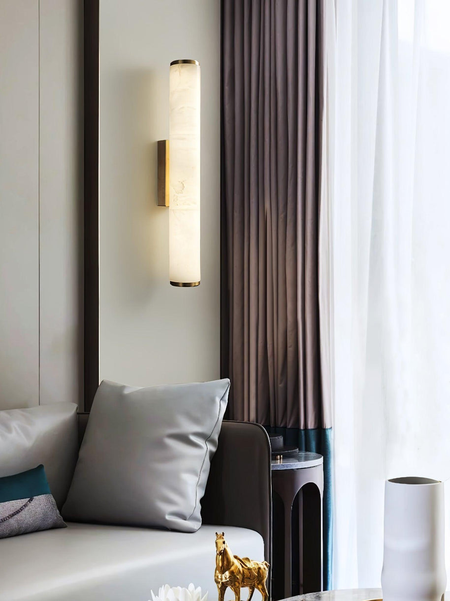 Wall lamp with a long pale veined alabaster tube and a metal bracket lit beside a curtain and a sofa.
