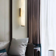 Wall lamp with a long pale veined alabaster tube and a metal bracket lit beside a curtain and a sofa.