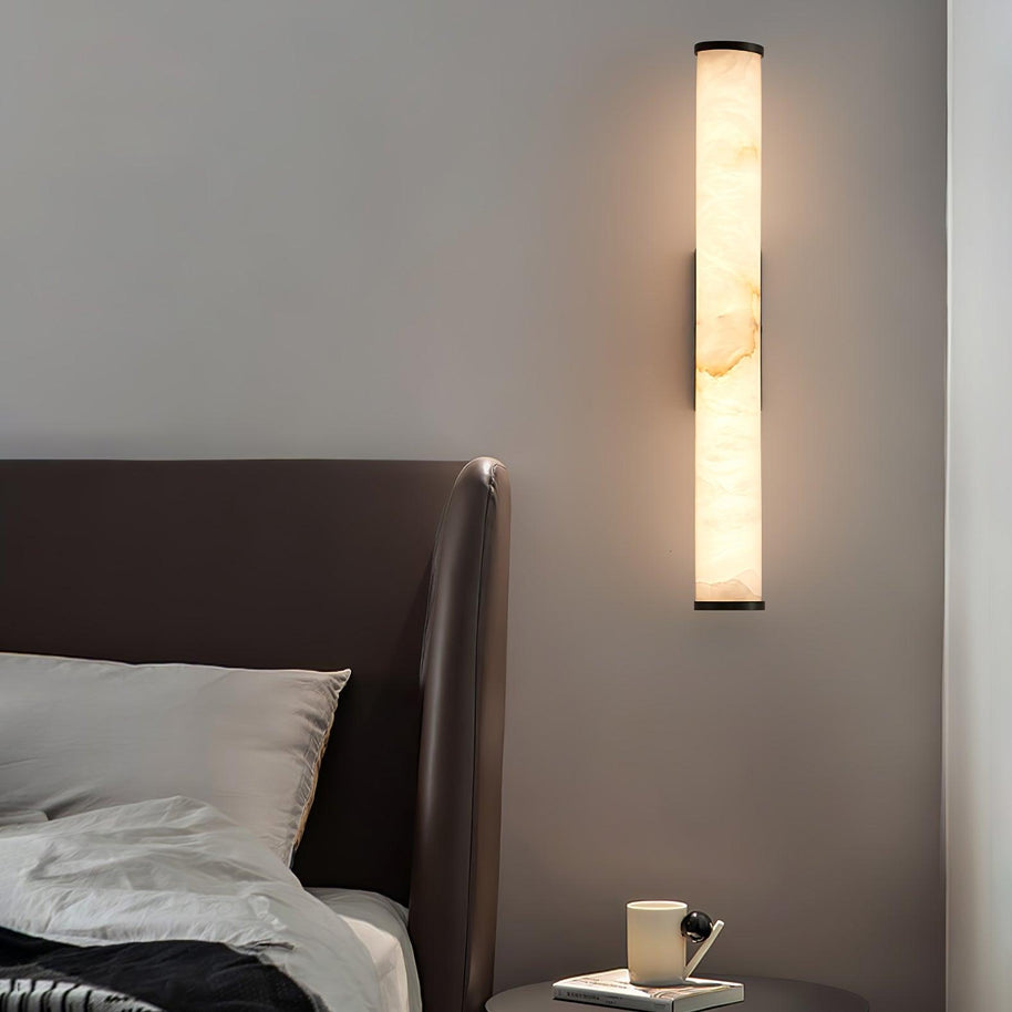 Wall lamp with a long pale veined alabaster tube and a black bracket lit beside a leather headboard.