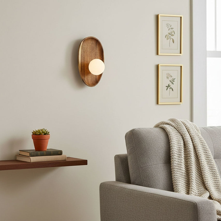 Wall lamp with a shaped walnut-coloured wooden backplate and a white glass globe on a wall beside two framed botanical prints.