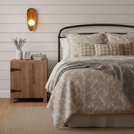 Wall lamp with a shaped walnut-coloured wooden backplate and a white glass globe lit on a white boarded wall above a bedside cabinet.