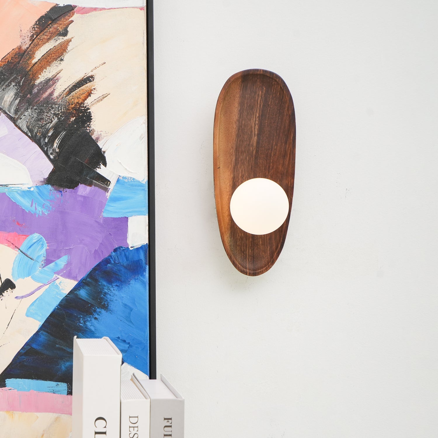 Wall lamp with a shaped walnut-coloured wooden backplate and a white glass globe lit beside a leaning framed print.