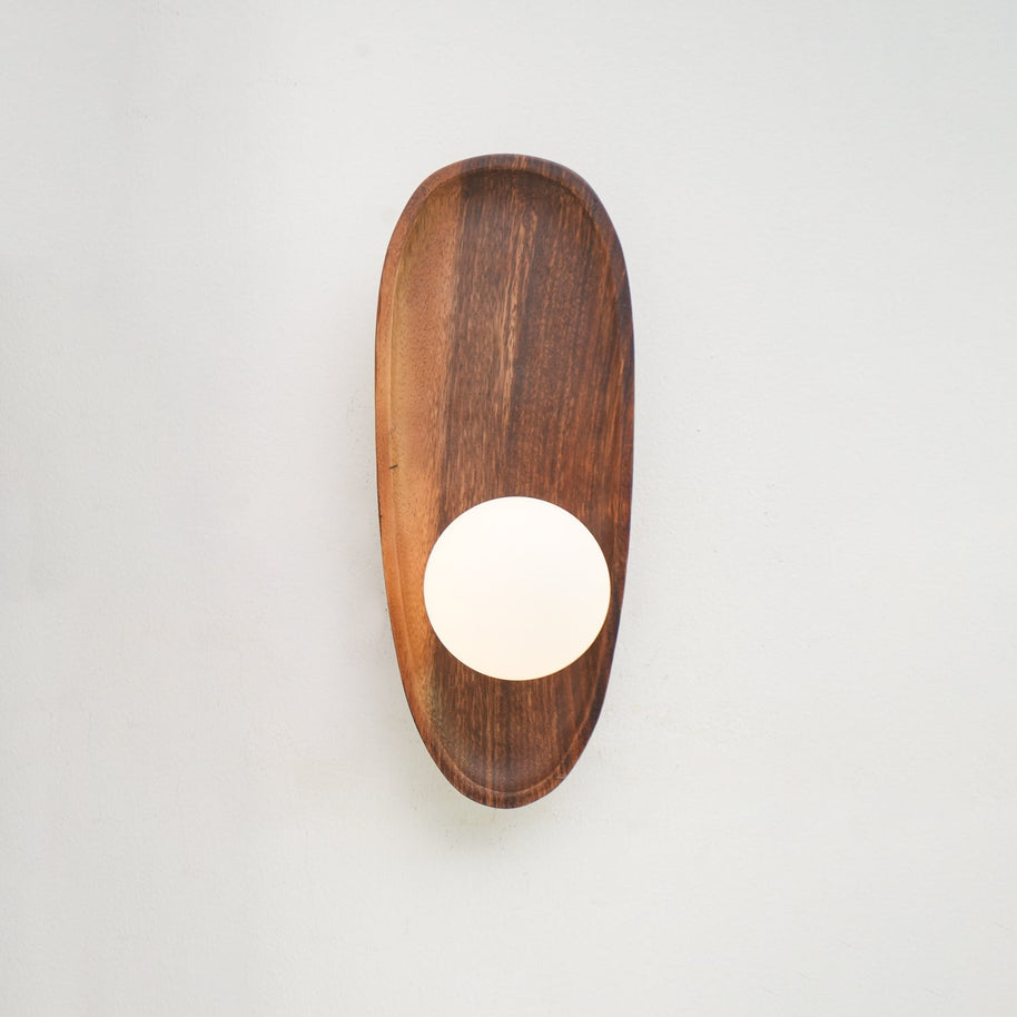 Wall lamp with a shaped walnut-coloured wooden backplate and a white glass globe lit, narrow upright shape, on a plain wall.