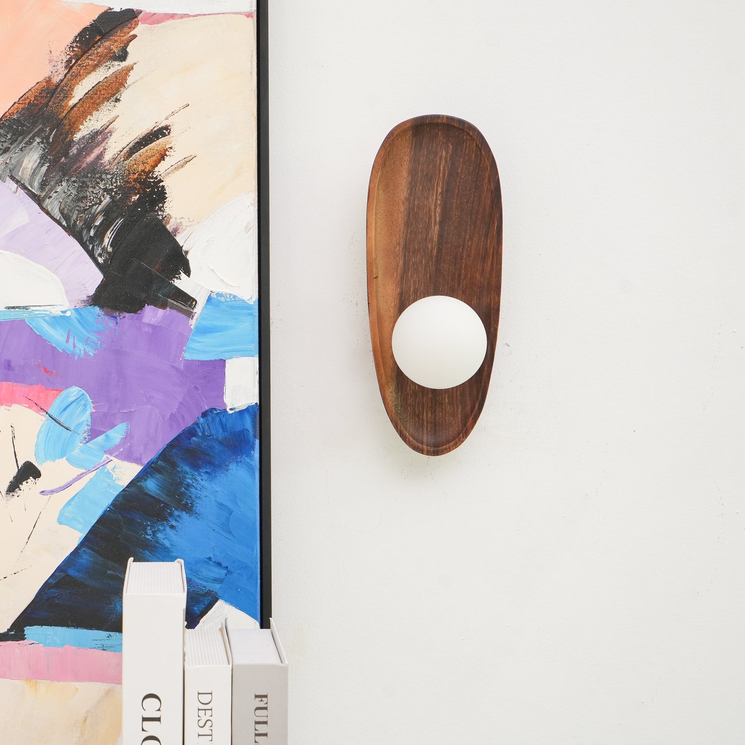 Wall lamp with a shaped walnut-coloured wooden backplate and a white glass globe beside a leaning framed print, unlit.