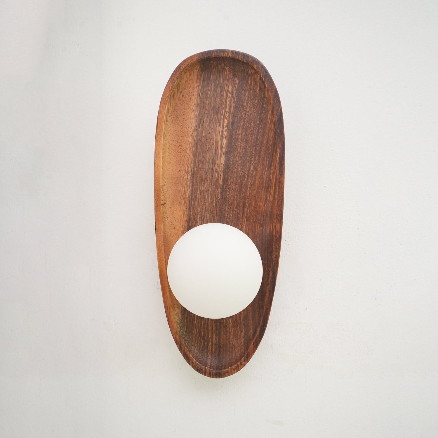 Wall lamp with a shaped walnut-coloured wooden backplate and a white glass globe lit, narrow upright shape, shown close on a plain wall.