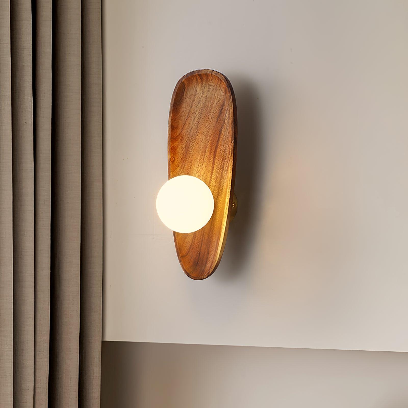 Wall lamp with a shaped walnut-coloured wooden backplate and a white glass globe lit beside a curtain and a wall panel.