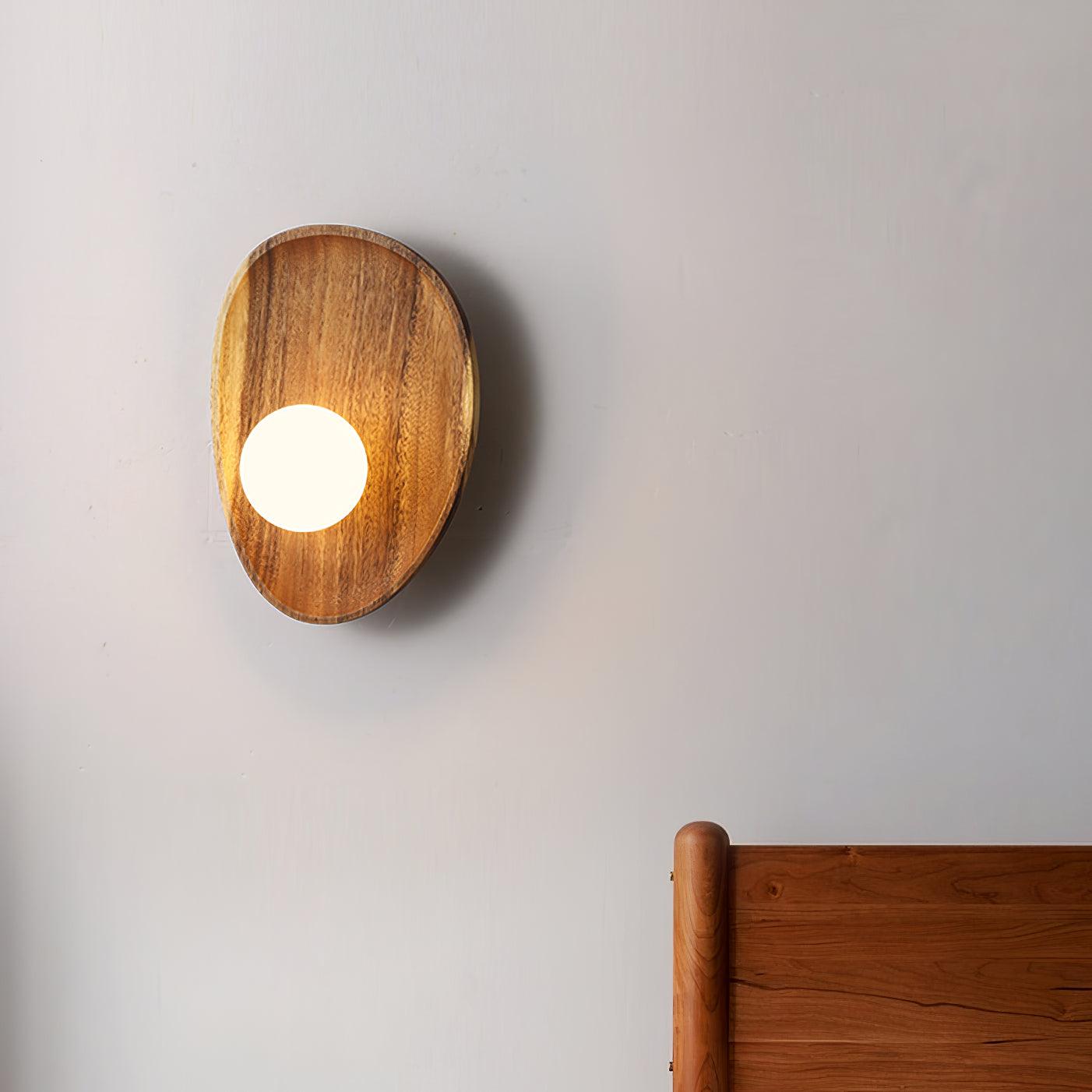 Wall lamp with a shaped walnut-coloured wooden backplate and a white glass globe lit, wider oval shape, above a wooden headboard.