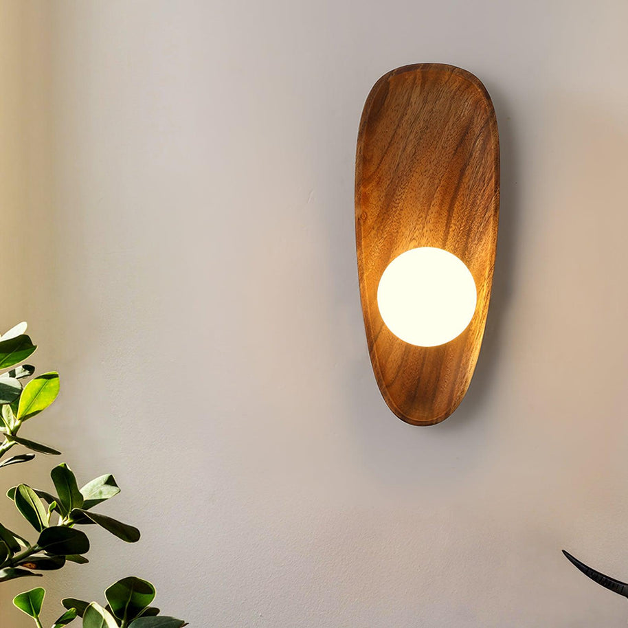 Wall lamp with a shaped walnut-coloured wooden backplate and a white glass globe lit, narrow upright shape, seen close beside a plant.